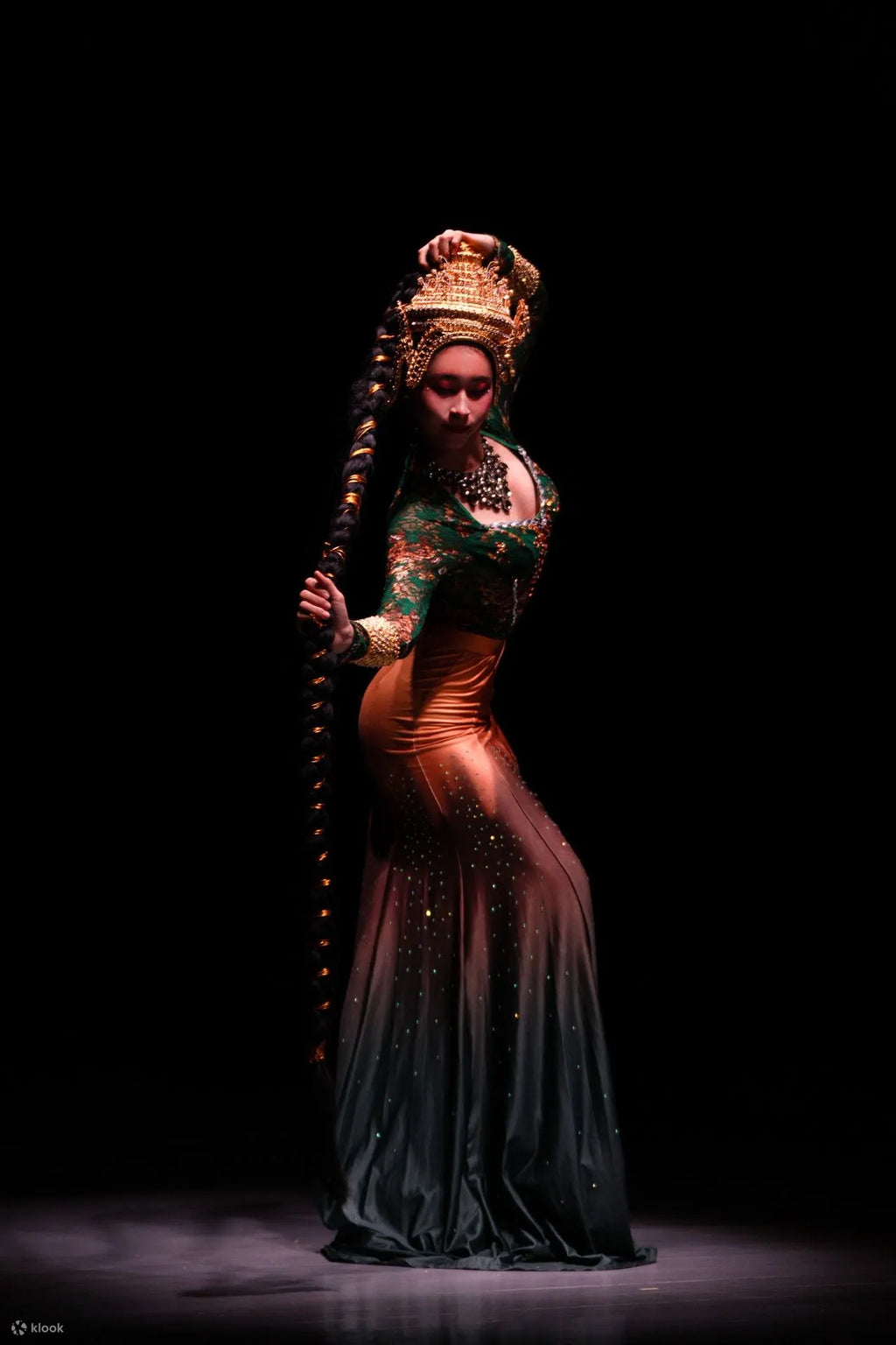 Dancer in traditional costume with intricate headpiece and long staff on a dark background
