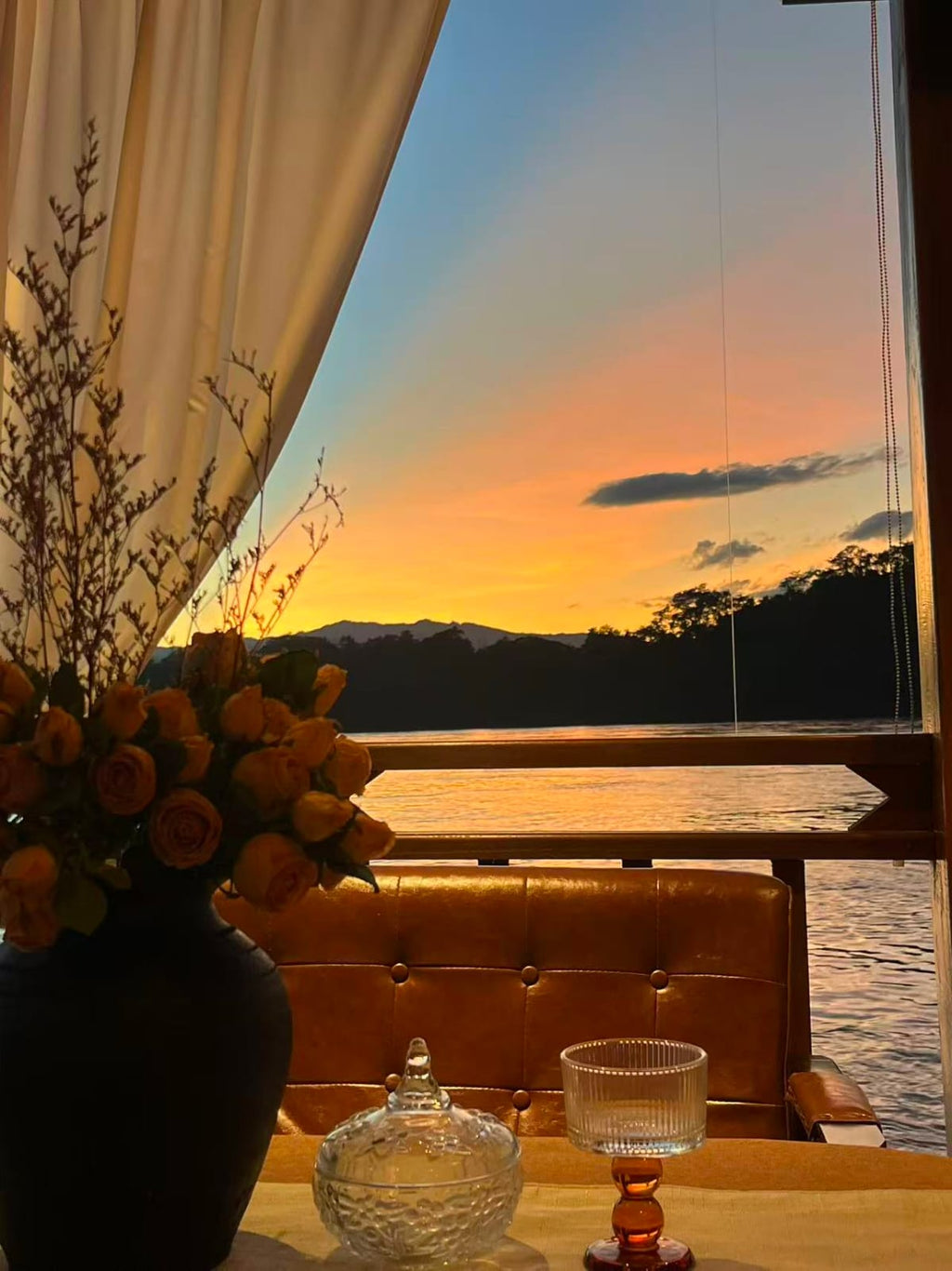 Sunset over a lake with a vase of flowers and a glass on a table.