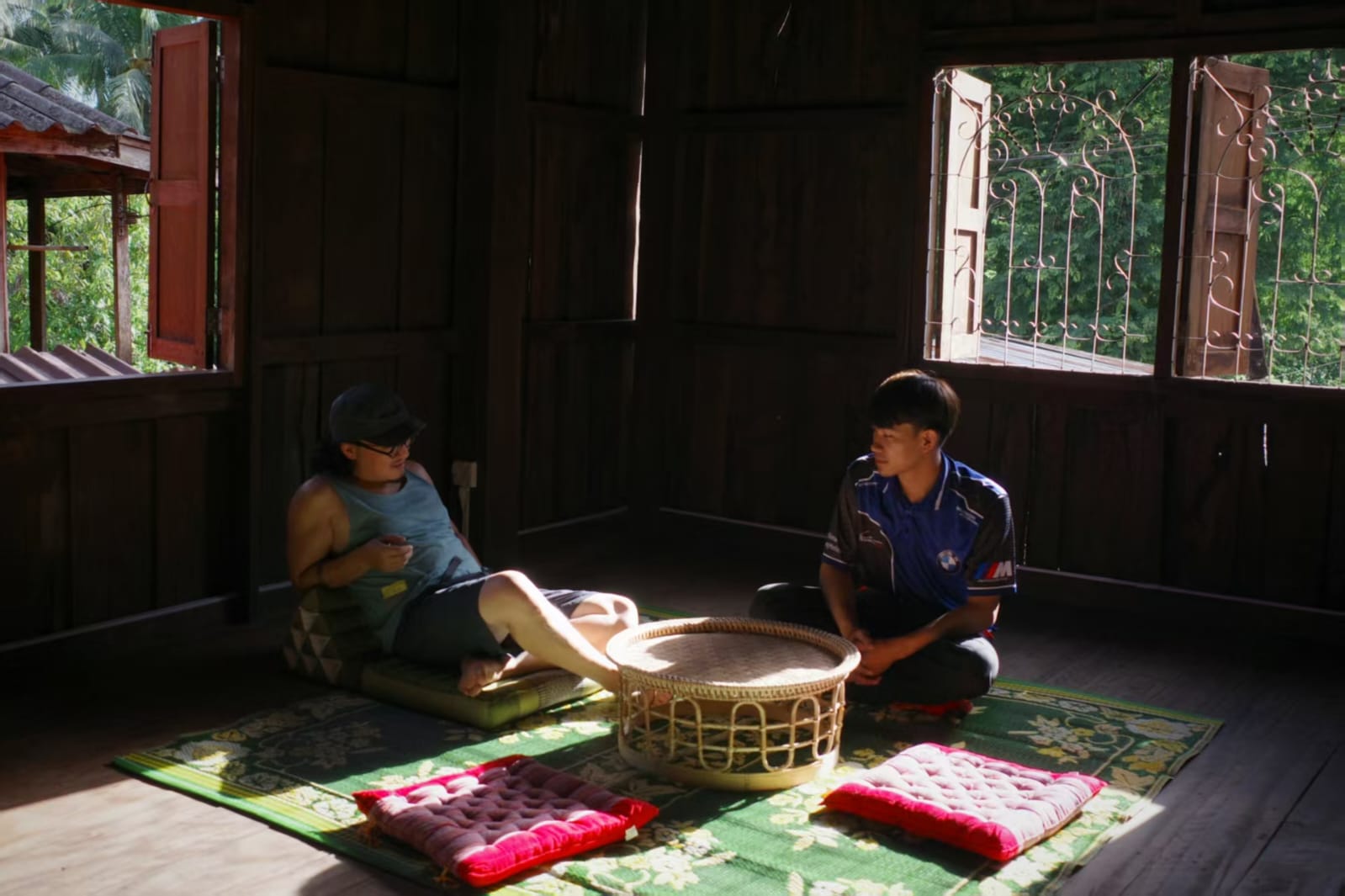 Two people sitting on mats in a room with a woven basket and colorful cushions. Xiangman Old House Café · Hiking Station